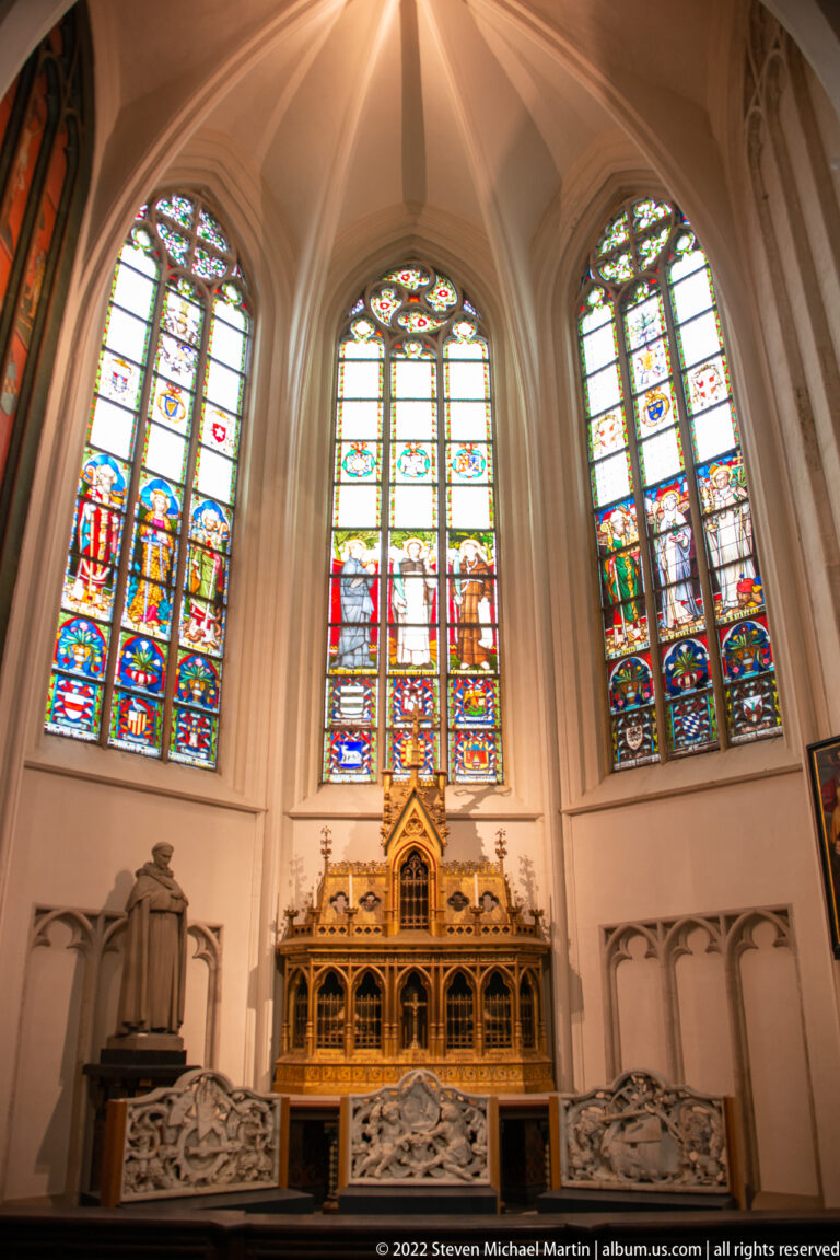 Three tall stained-glass windows beneath pale rib vaults, with a gilded Gothic-style shrine and stone reliefs below.