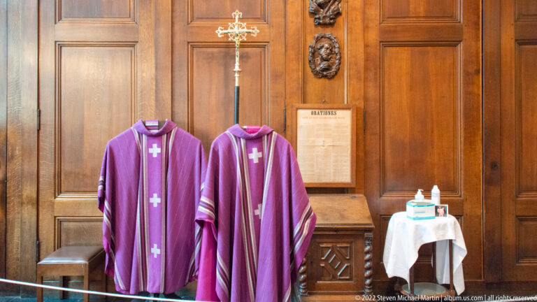 Two purple chasubles with pale crosses hanging before wood paneling, with a tall cross and small white-covered table nearby.