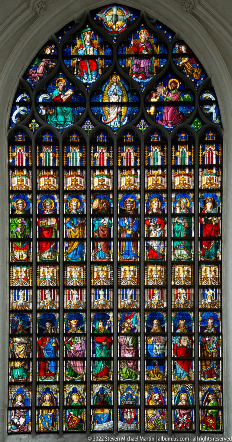 Large pointed stained-glass window divided into many narrative scenes, heraldic shields, and figures beneath dark tracery.