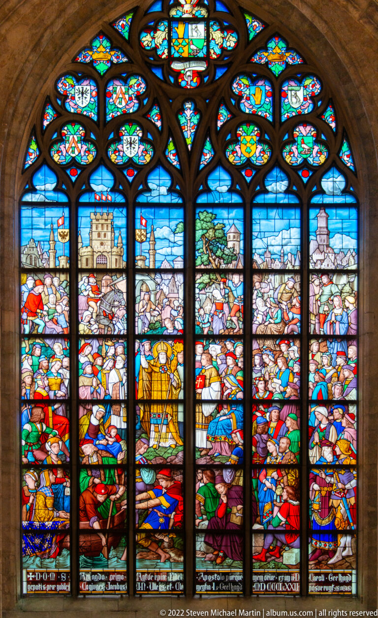 Broad stained-glass window with crowded historical scenes, heraldic emblems in the tracery, and inscriptions along the bottom.