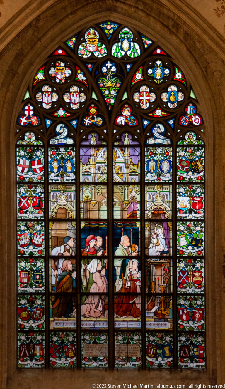 Pointed stained-glass window with three main narrative panels surrounded by dense heraldic, floral, and geometric borders.