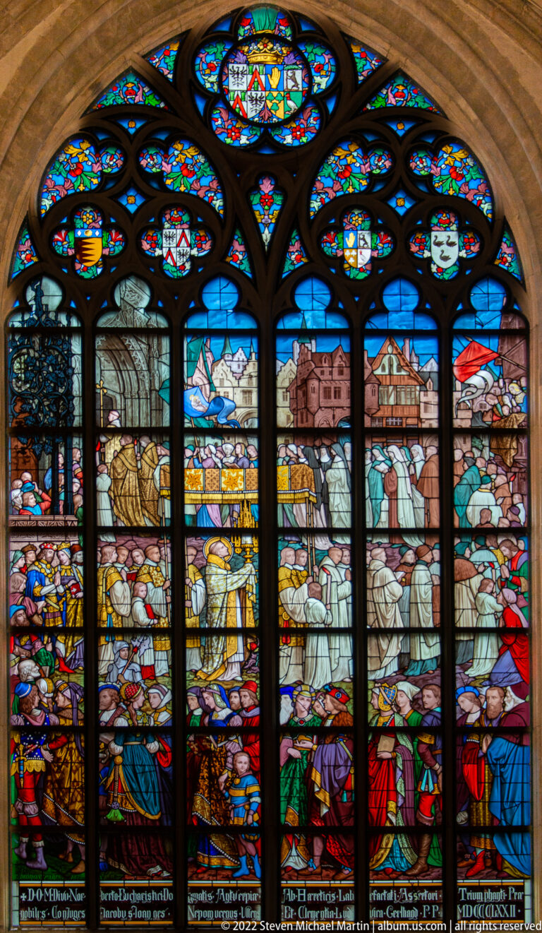 Multi-lancet stained-glass window showing clergy, nobles, attendants, architecture, landscapes, heraldry, and inscriptions.