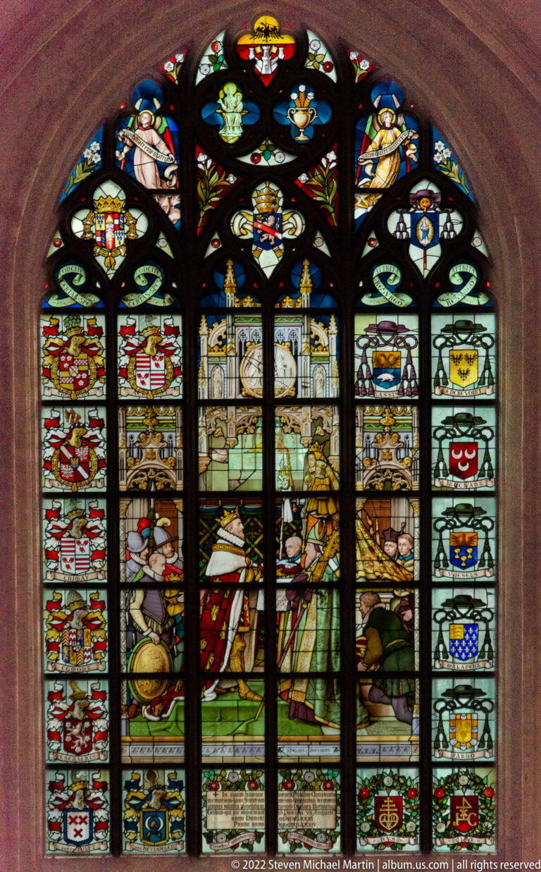 Pointed stained-glass window with central standing figures, numerous coats of arms, and smaller narrative panels around them.