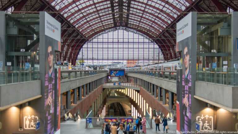 View down into Antwerp-Central Station’s lower levels with escalators, passengers, platforms, and red roof structure above.