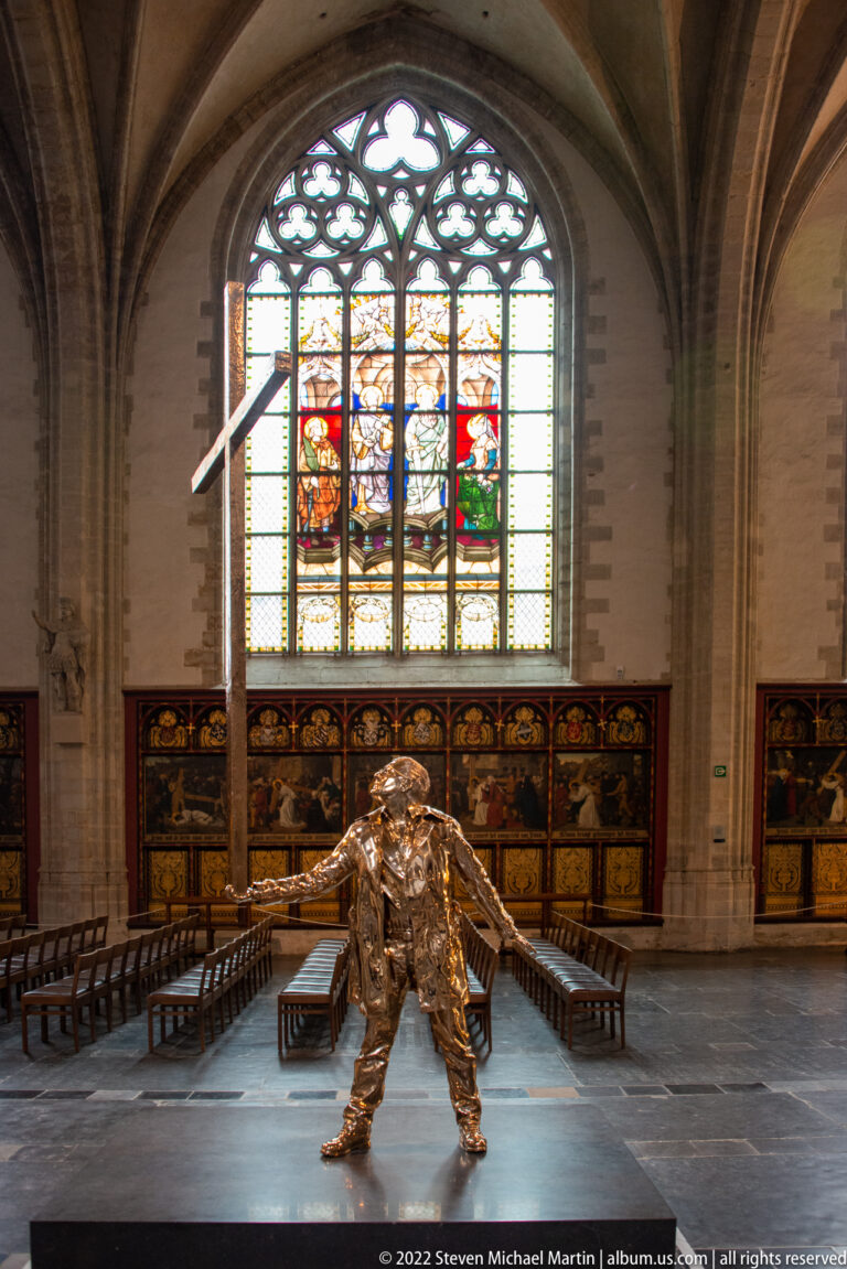 Bronze-colored crouching human sculpture centered in a cathedral aisle, with choir stalls and stained glass behind.