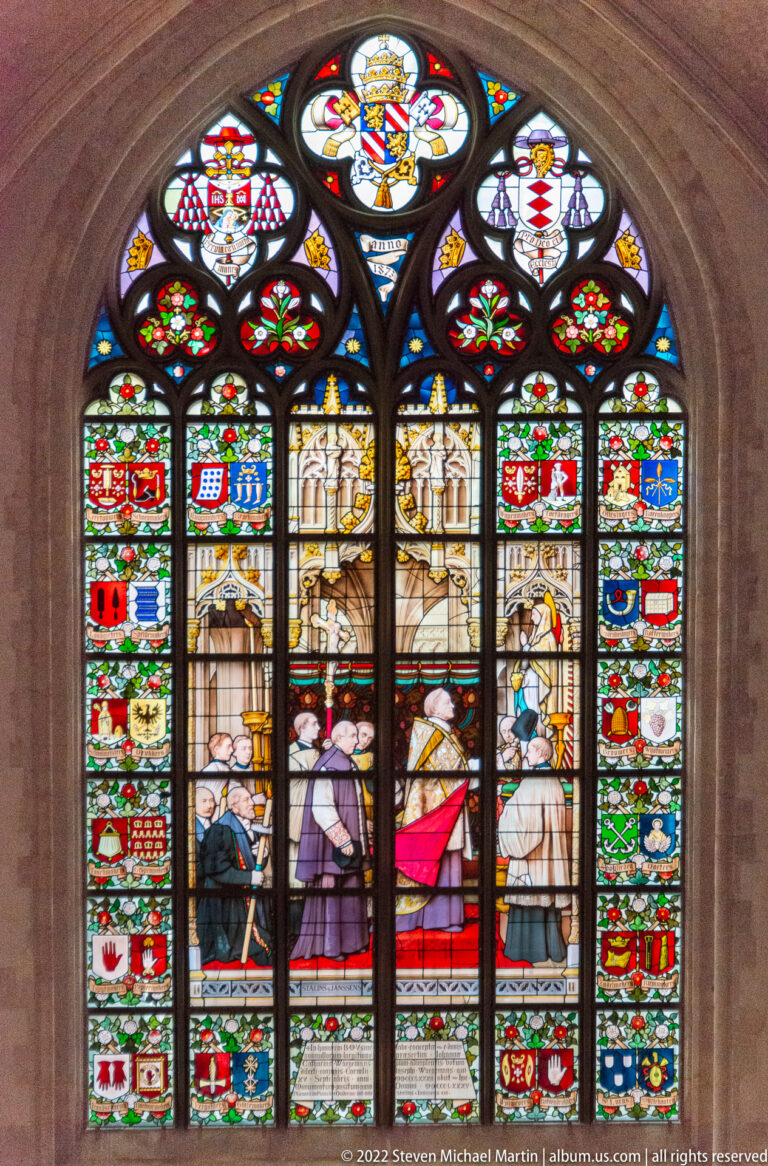 Stained-glass window with standing and kneeling figures, heraldic shields along both sides, and a sacred figure in the top roundel.