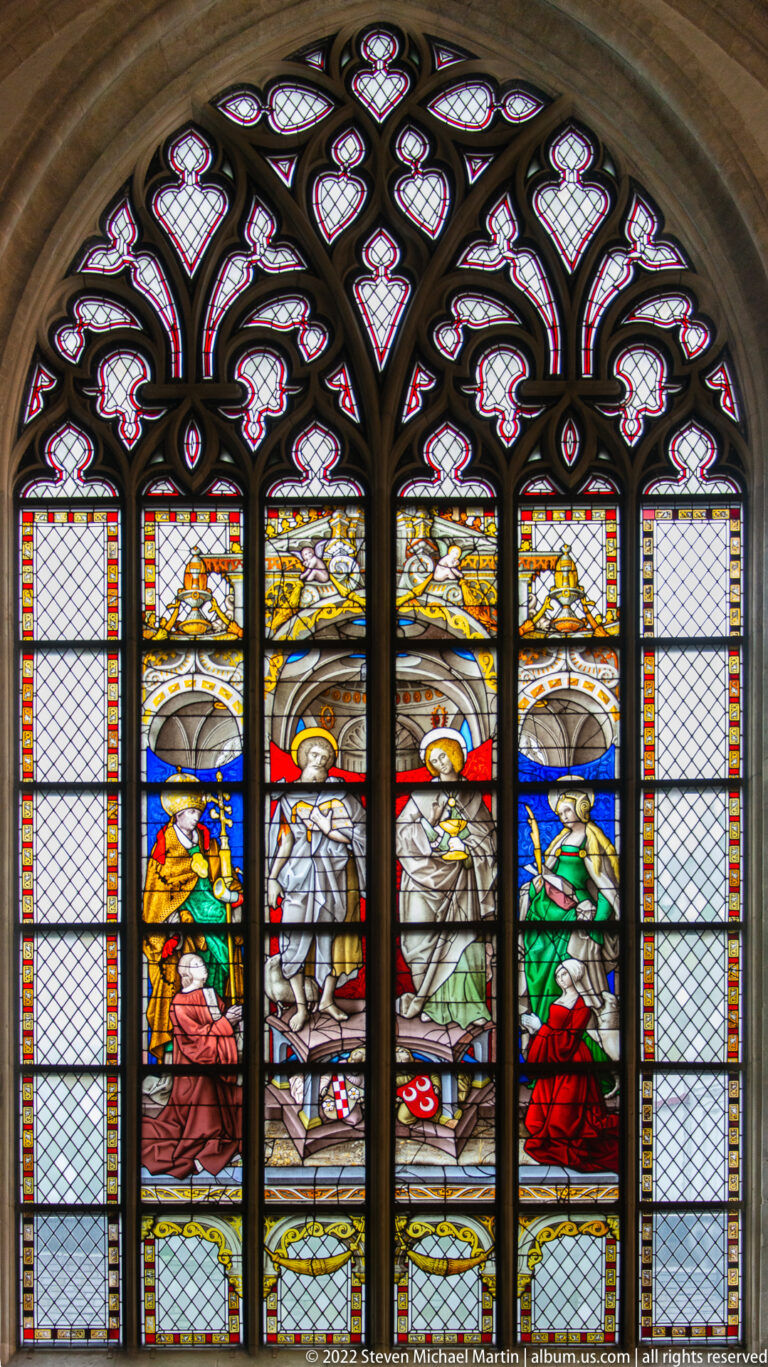 Four robed figures in colored stained glass beneath pale sweeping forms and pink-and-white ornamental Gothic tracery.