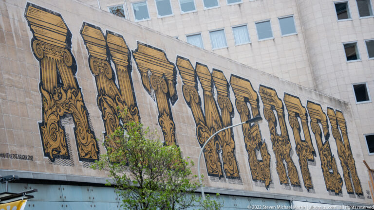 Large wall mural spelling ANTWERPEN in ornate black-and-gold patterned letters, with a modern building rising behind.
