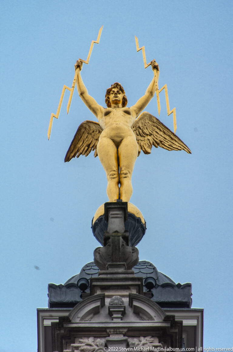 Winged putto standing atop a dark pediment and raising golden lightning-bolt forms against a blue sky.