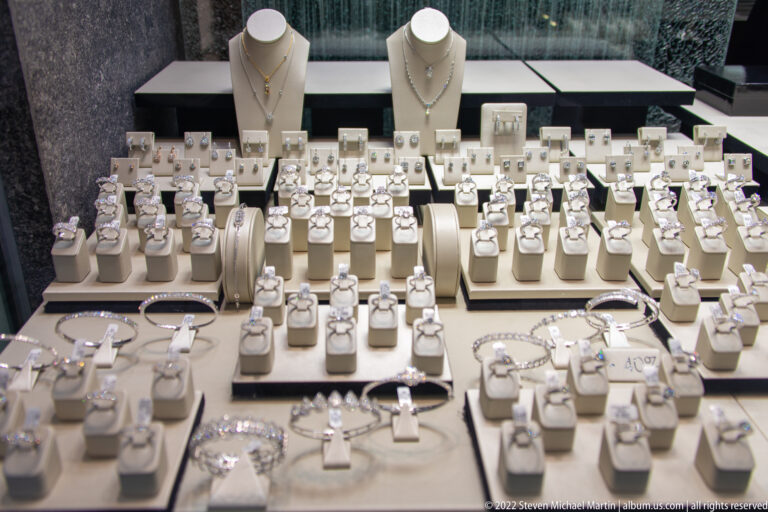 Jewelry shop display densely arranged with rings, bracelets, necklaces, and earrings on pale stands.