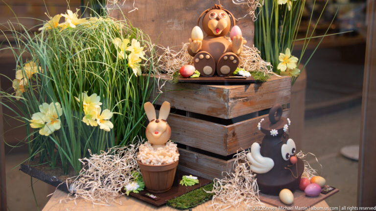Easter chocolate display with cartoon-like figures, rabbit, bird, pastel eggs, flowers, grass, straw, and wooden crates.