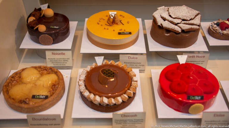 Display case of glossy round cakes in chocolate, caramel, yellow, and red finishes with individual flavor labels below.