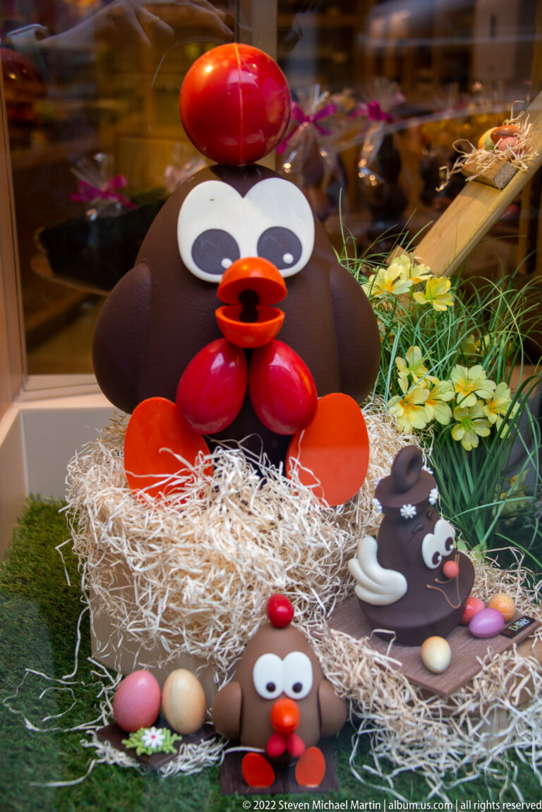 Large cartoon chocolate bird in a straw nest with smaller bird figures, pastel eggs, greenery, and spring flowers.