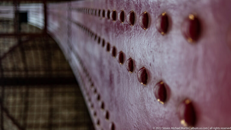 Close-up of rivets on a red-painted steel beam, with the station roof structure blurred behind.
