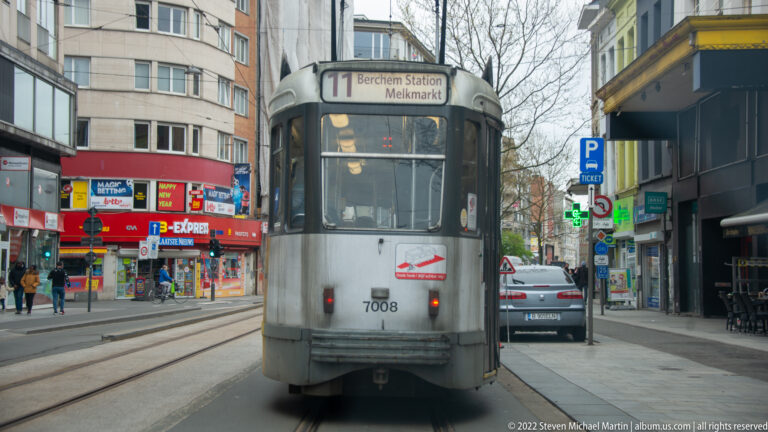 Silver Antwerp tram number 7008 on route 11 toward Berchem Station, traveling along tracks through a commercial street.