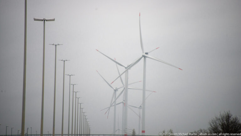 Wind turbines fading into heavy gray fog beside a roadway lined with tall streetlights and utility poles.