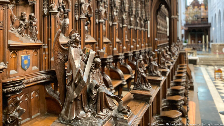Carved wooden angel beside a seated figure at the end of ornate choir stalls lined with saints and Gothic canopies.