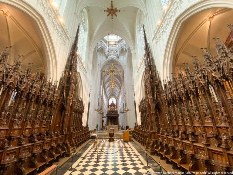 Wide symmetrical cathedral choir with richly carved wooden stalls, pale soaring vaults, and black-and-white patterned floor.