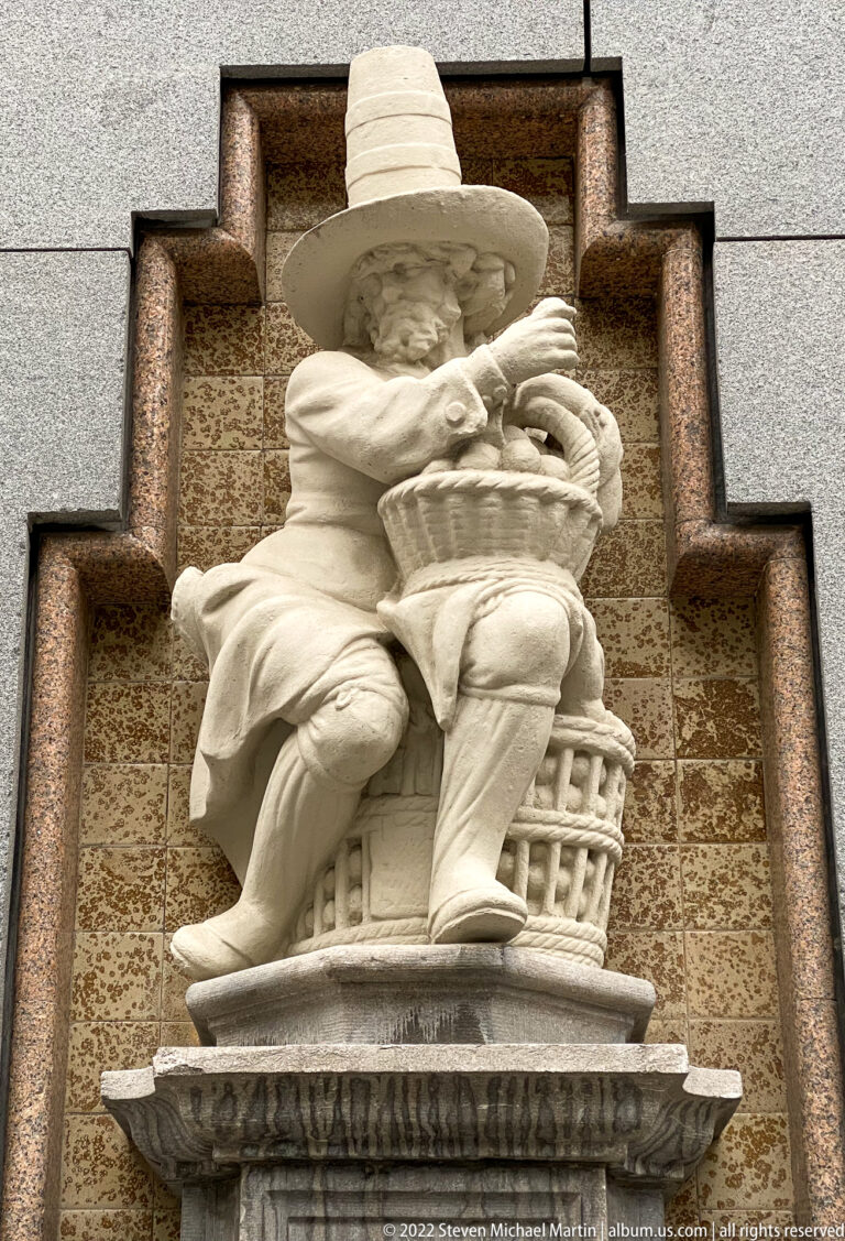 Seated pale stone figure in a broad hat holding a small object, mounted against patterned masonry within a dark stepped frame.