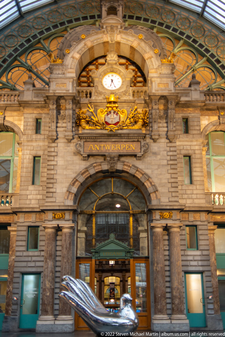 Polished metal sculpture before the ornate ANTWERPEN facade, clock, and gilded crest inside Antwerp-Central Station.