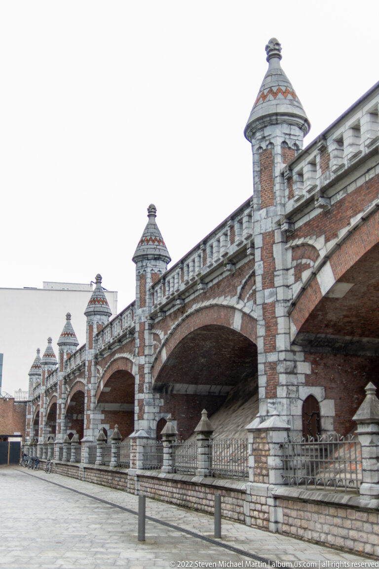 Broad brick railway arches and banded stone piers receding beside an open paved area in central Antwerp.