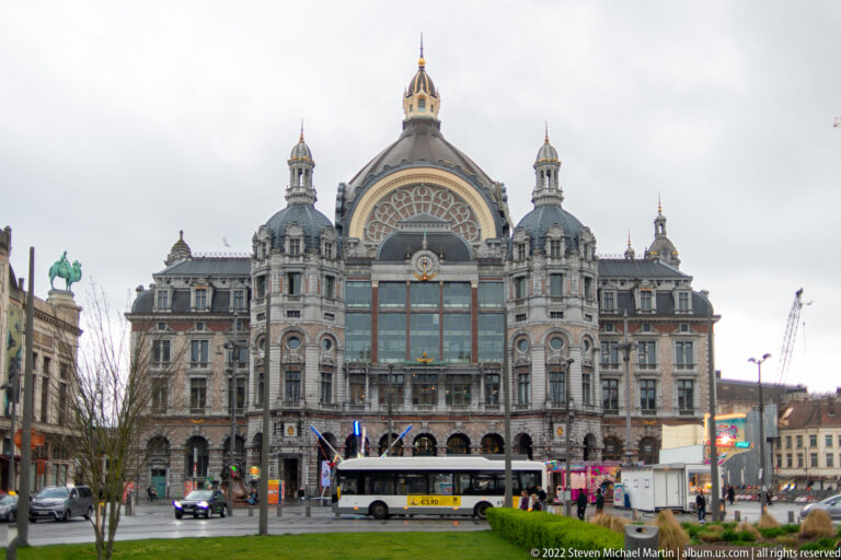 Front exterior of Antwerp-Central Station with central arched window, towers, domes, buses, and street activity.