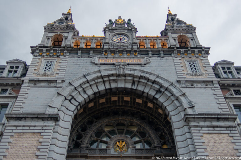 Upward view of Antwerp-Central Station’s massive stone entrance arch, fan window, clock, and rooftop ornament.