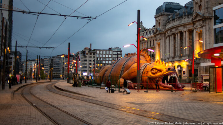 Large illuminated dragon with open jaws beside curving tram tracks and city buildings at dusk in Antwerp.