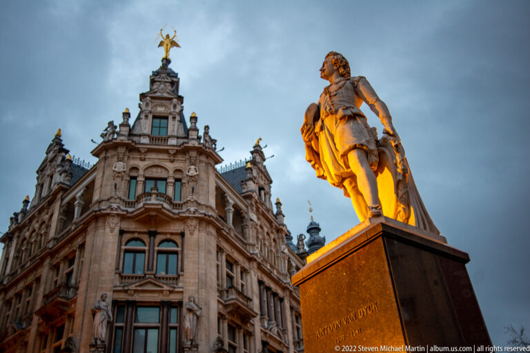 Illuminated statue of Anthony van Dyck on a dark pedestal before an ornate building on Antwerp’s Meir.
