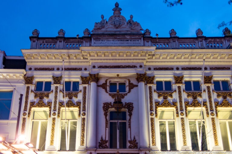 Illuminated ornate white stone facade with tall windows, gilded decoration, and sculpted roofline against a blue evening sky.