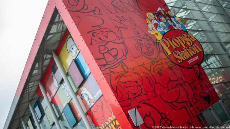 Red Plopsa Station facade with cartoon graphics and round entrance sign beneath a glass canopy in Antwerp.