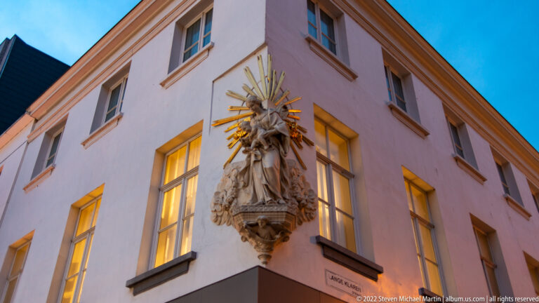 Carved religious figure surrounded by gilded sunburst rays on the corner of a pale building at dusk.