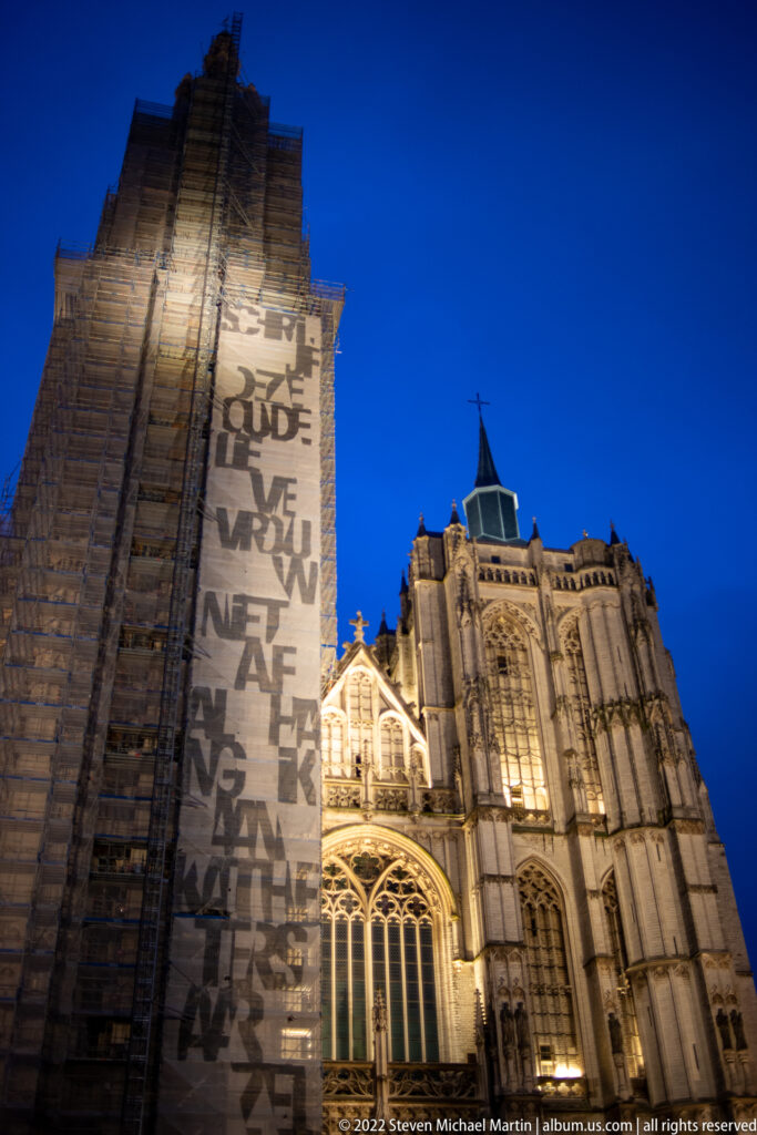 Night view of Antwerp Cathedral’s Gothic exterior beside a wall covered by a large projected text installation.
