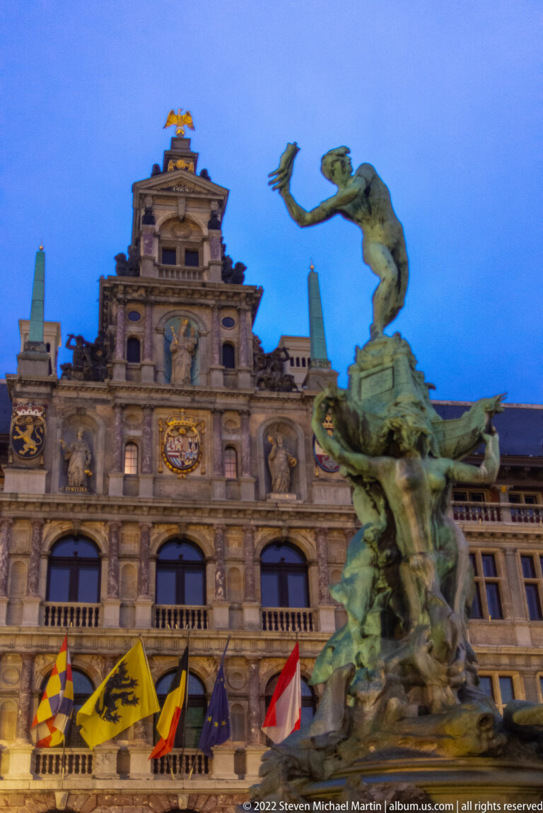Brabo Fountain in the foreground with Antwerp City Hall and flags behind it at dusk.