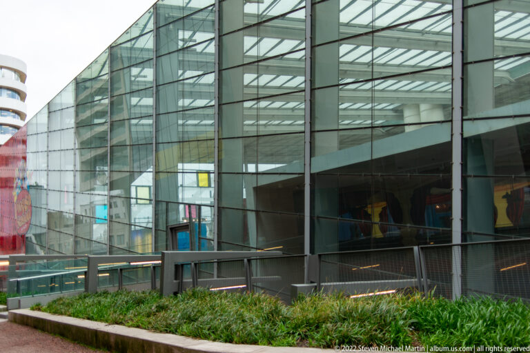 Glass-and-steel concourse with gridded roof, transparent walls, and a planted strip at the base.