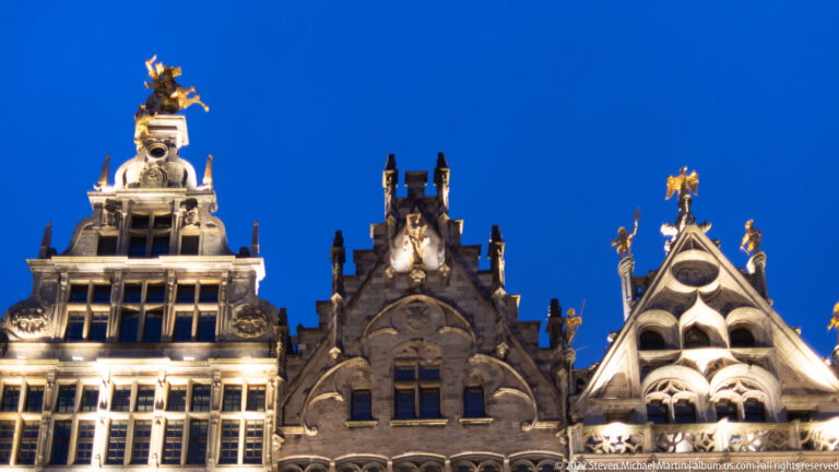 Close view of three ornate Grote Markt guildhouse gables with gilded rooftop figures against a blue evening sky.
