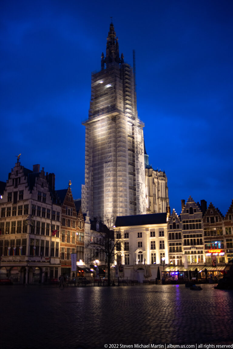 Antwerp Cathedral tower wrapped in scaffolding above illuminated Grote Markt buildings at night.