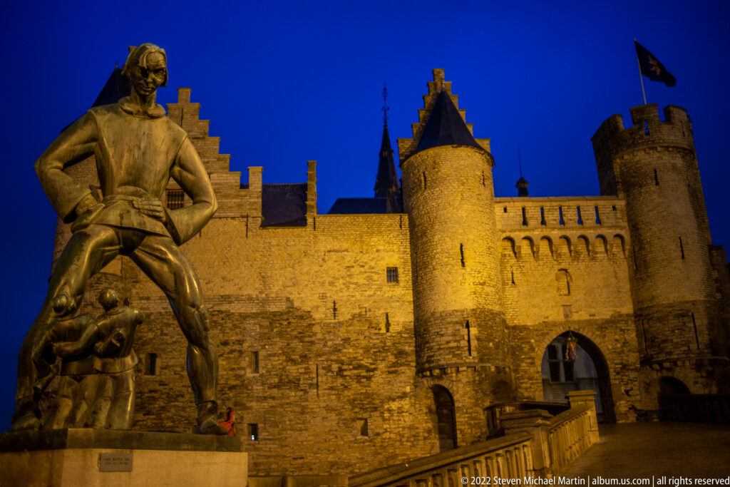Warmly lit Het Steen gate and round towers behind the large Lange Wapper statue at night.