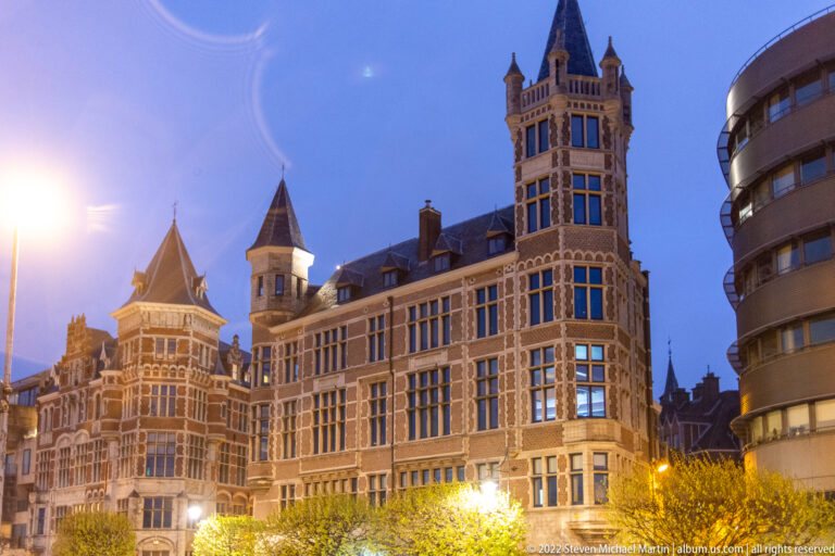 Ornate historic building with mullioned windows and a tall corner turret beside a rounded modern building in Antwerp.