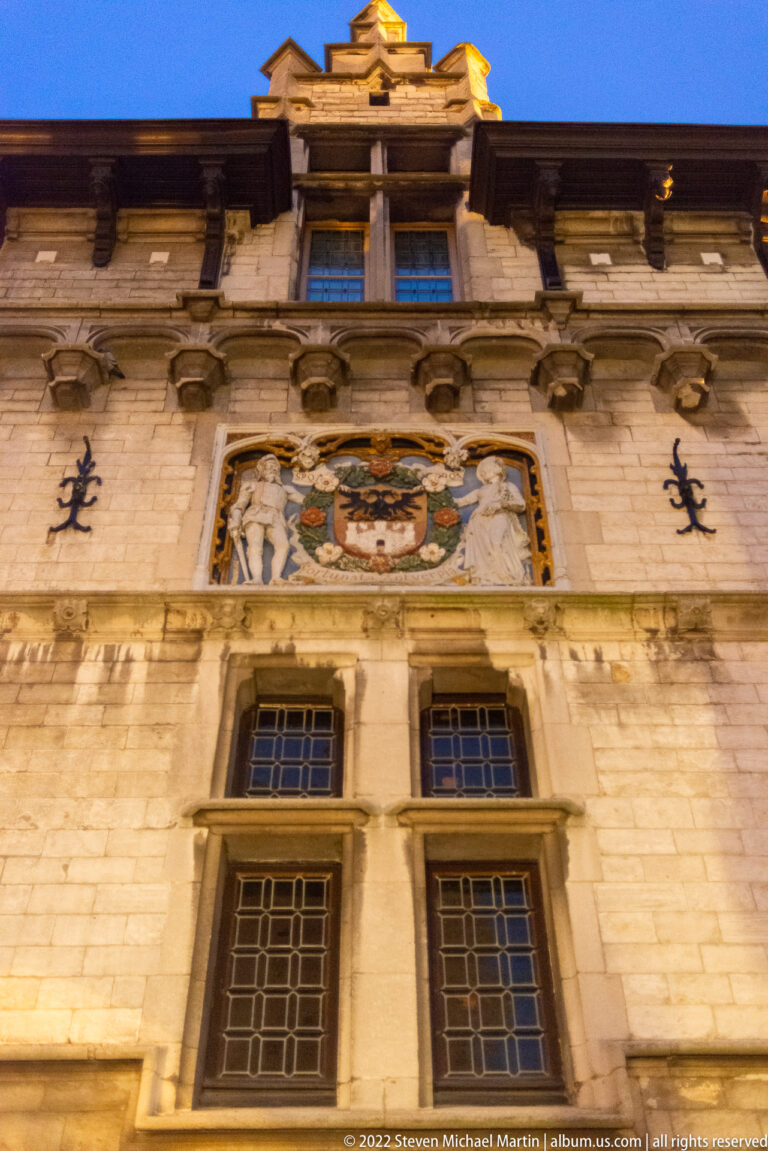 Painted coat of arms set within carved pale-stone ornament above windows on a historic Antwerp building.