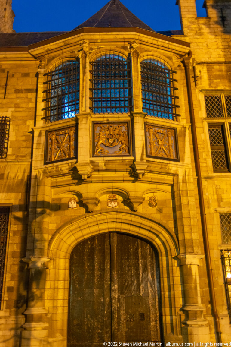 Large closed arched gateway beneath gridded windows and carved stone panels, illuminated at night in Antwerp.