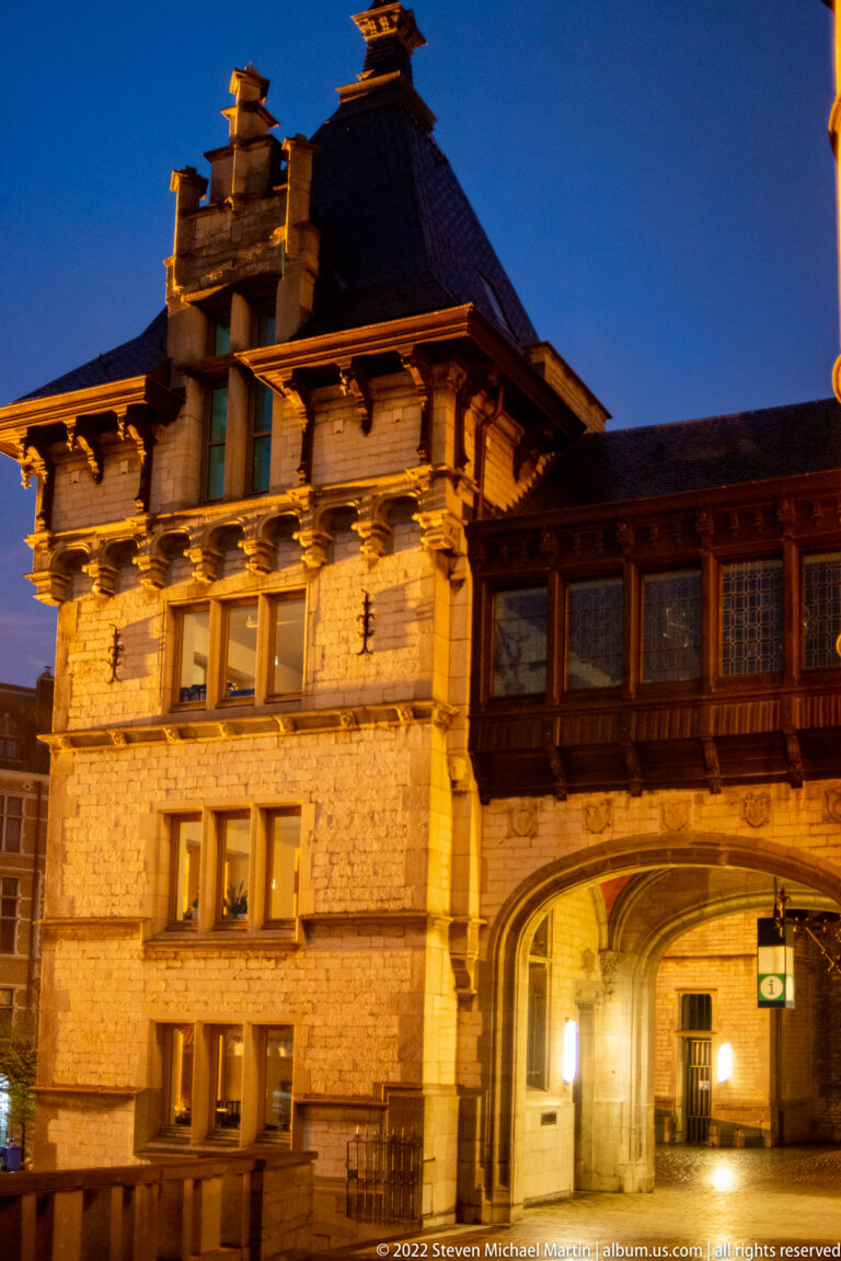 Historic masonry passage with a projecting upper story, small windows, and a dark roof, illuminated at night.