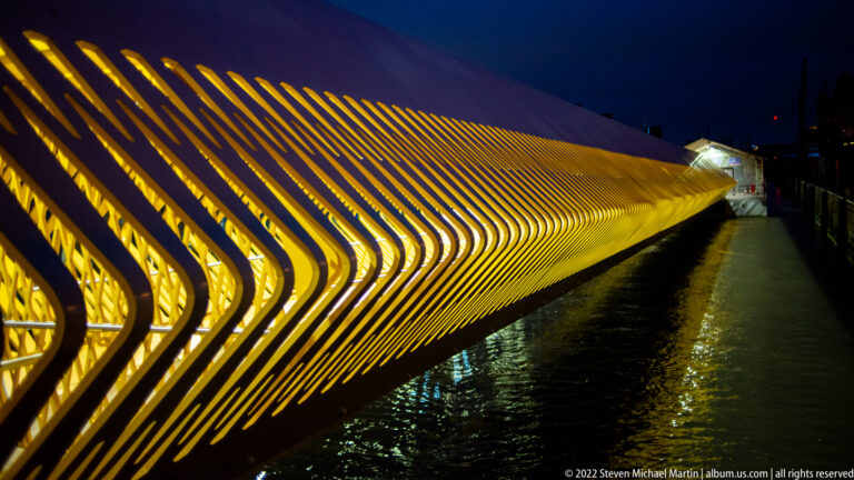 Long illuminated pedestrian bridge with closely spaced diagonal ribs forming a glowing geometric screen at night.