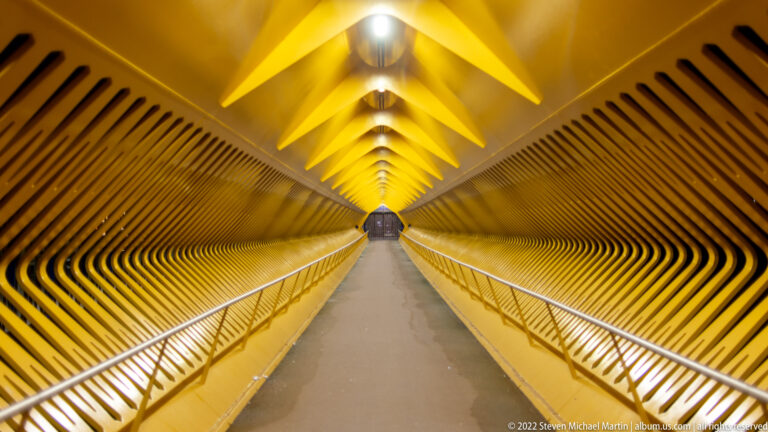 Symmetrical interior of a yellow illuminated pedestrian bridge with repeating ribs, handrails, and a distant opening.