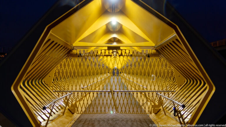 Head-on view toward a metal lattice screen at the end of a yellow ribbed pedestrian bridge.