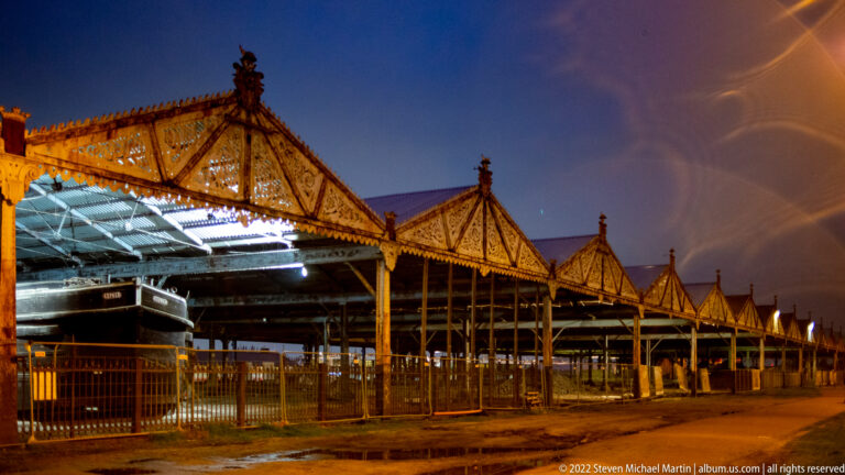 Long historic iron-and-glass railway shed with repeated triangular roof bays, lit from within at night.