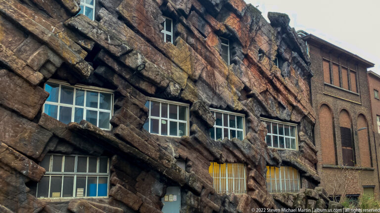 Artificial cliff-like rockwork façade with rectangular windows beside a brick building in Antwerp.