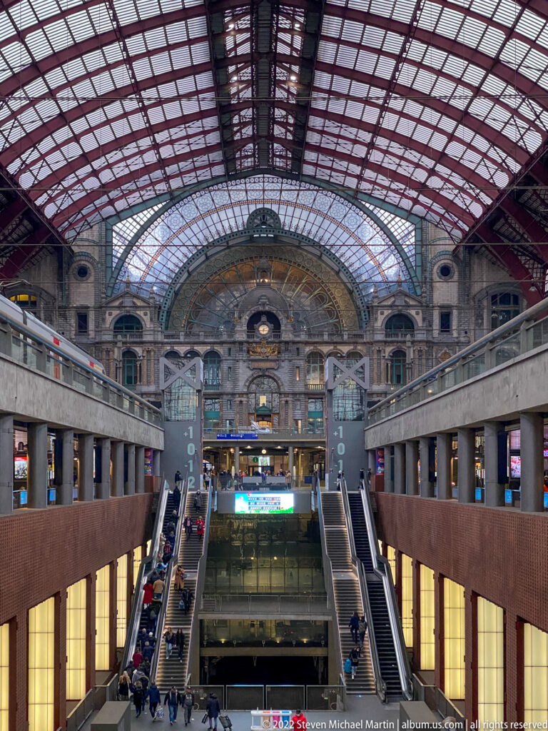 Symmetrical interior of Antwerpen-Centraal beneath the vast glass train shed, with escalators and lower passenger levels.