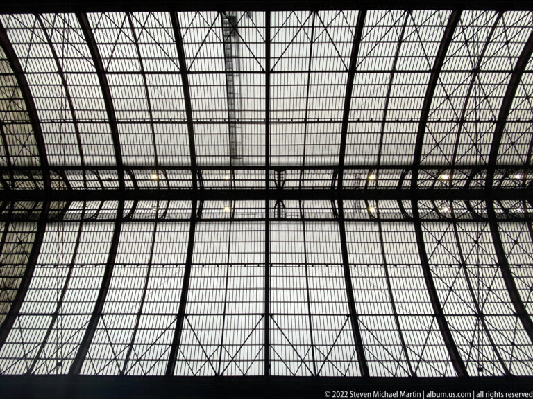 Upward view of Antwerpen-Centraal’s iron-and-glass train-shed roof with dense trusses and rectangular glazing.