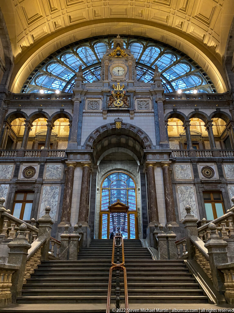 Grand staircase inside Antwerpen-Centraal rising toward an arched window beneath richly decorated stonework.